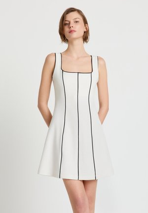 Frau trägt ein ärmelloses weißes Kleid mit schwarzen vertikalen Streifen und einem quadratischen Ausschnitt, steht vor einem schlichten hellen Hintergrund.