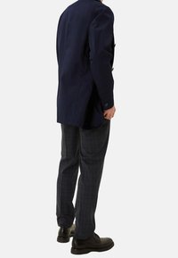 Hombre con un blazer azul marino, pantalones de cuadros grises y zapatos de vestir negros, de pie contra un fondo liso, visto desde detrás.