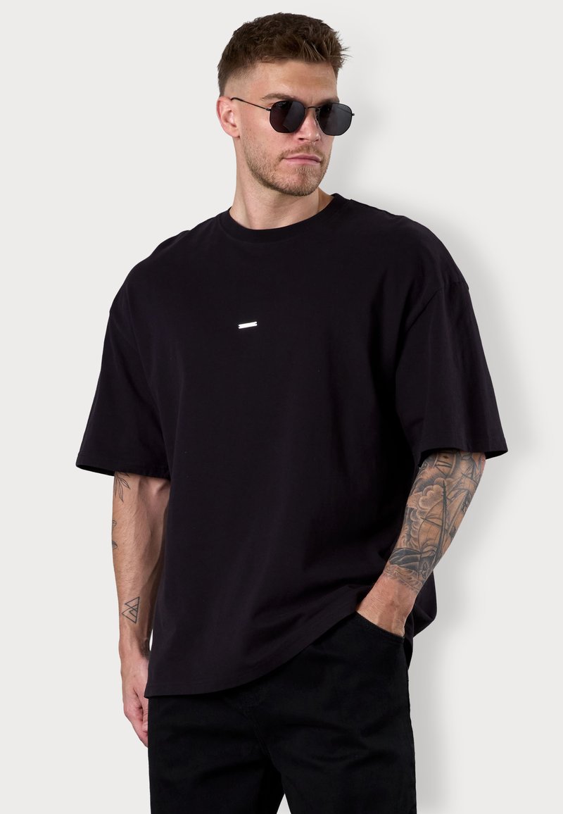 Schwarzes, übergroßes T-Shirt aus Baumwolle mit kurzen Ärmeln und einem kleinen reflektierenden Detail auf der Brust. Getragen mit schwarzen Hosen.