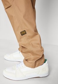 Beige cargobukser med synlige sømmer og G-Star-merke, kombinert med hvite sneakers med perforerte detaljer og grønne aksenter.