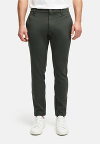Pantalones ajustados de color verde oscuro hechos de una tela suave, con cierre de botón, trabillas para cinturón y bolsillos laterales, combinados con zapatillas blancas.