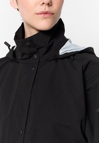 Vrouw in een zwarte waterdichte jas met hoge kraag en drukknoopsluiting, gedeeltelijk opengeritst met een capuchon die een lichtgrijze voering toont.