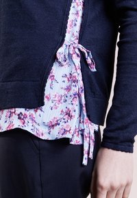 Marinblått stickat cardigan över en ljusblå blommig blus med en knuten detalj, med en texturerad stof och utsvängd nederkant.