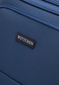 Tmavě modrá taška z látky s texturovaným povrchem, se černým obdélníkovým štítkem s raženým slovem "WITTCHEN". Viditelné detaily zipu.