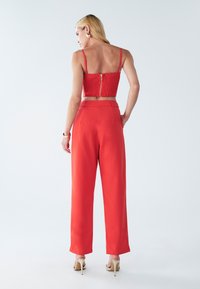 Crveni skraćeni smotan top s patentnim zatvaračem na leđima, uparen s hlačama širokog kroja viskog struka. Glatka tkanina, minimalni detalji i sandale s remenčićima.