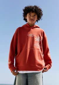 Rust-colored hoodie with palm tree design, "LOS ANGELES CALIFORNIA" text, kangaroo pocket, and ribbed cuffs. Worn over a white shirt.