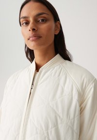 Veste bombardier crème avec une texture matelassée, un col montant et une fermeture éclair à l'avant. Tissu lisse avec de subtils motifs en losange sur toute la surface.