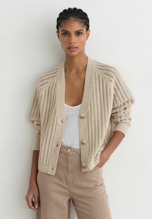Vrouw draagt beige gebreid vest met knopen over wit topje en lichtbruine broek met hoge taille, staand tegen een witte muur.