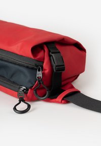 Rode en zwarte rol-tas met een zwarte verstelbare schouderband, twee ritsen met ronde trekkoorden en een zwarte plastic sluiting met een gesp.