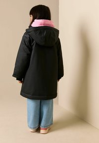 Juodos spalvos išorinis paltas su gobtuvu, rausvas vidinis pamušalas, derinamas su šviesiai mėlynomis plačiomis džinsų kelnėmis ir smėlio spalvos batais. Minimalistinis dizainas, lygus paviršius.