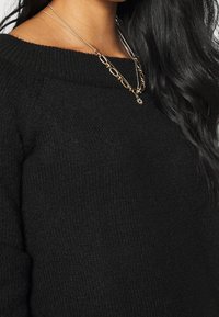 Maglione nero lavorato a maglia con scollatura ampia, caratterizzato da un tessuto dalla texture interessante. Collane in catena d'oro sovrapposte con un pendente completano il look.