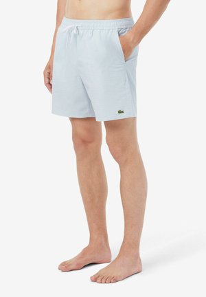 Homme portant un short à carreaux bleu clair avec des poches latérales et une taille à cordon, debout pieds nus sur un fond blanc.