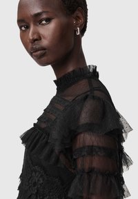 Svart genomskinlig klänning med volangärmar, texturerat tyg och hög krage. Designen har spetsdetaljer och fack med volanger.