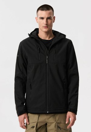 Mężczyzna ubrany w czarną kurtkę z kapturem zapinaną na zamek oraz beżowe spodnie typu cargo, stojący na jasnym, jednolitym tle.