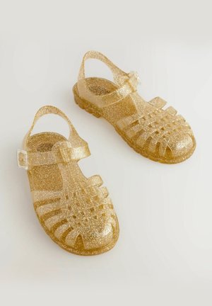 Glitzernde goldene Gelly-Sandalen mit geschlossener Spitze, offenen Seiten und verstellbarem Knöchelriemen. Strukturiert Oberfläche und flexibles Material.