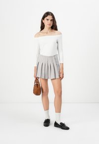 Top bianco con spalle scoperte e maniche lunghe abbinato a una mini gonna plissettata grigia. La modella tiene in mano una borsa marrone, indossando calze bianche e scarpe nere.