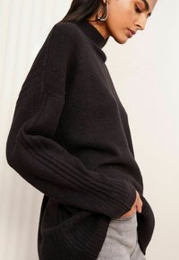 Schwarzer Strickpullover mit hohem Kragen, gerippten Ärmeln und lockerer Passform, kombiniert mit hellgrauen Jeans. Weicher Stoff und dezente Details.