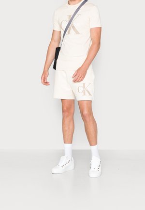 Beige Baumwoll-T-Shirt mit "CK"-Logo, kombiniert mit passender Shorts. Weiße Sneakers und Socken runden das Outfit ab, ergänzt durch eine schwarze Schultertasche.