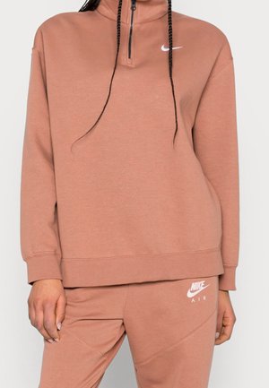 Osoba ubrana w brązowy dres Nike Air z bluzą na zamek i dopasowanymi spodniami, z białymi logo Nike na klatce piersiowej i udzie.