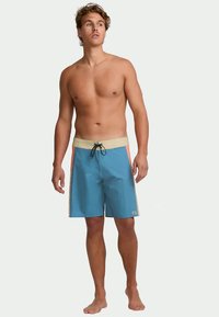 Αμετακίνητος νεαρός άνδρας χωρίς μπλούζα, φορώντας μπλε μαγιό με μπεζ ζωνάκι και πορτοκαλί ρίγες στο πλάι, στέκεται ξυπόλυτος μπροστά από έναν ουδέτερο φόντο.