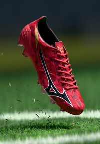 Sarkanais futbola apavs ar ģeometriskiem rakstiem, melniem akcentiem un vairākām naglām. Apavam ir elegants dizains un teksturēta virsma.