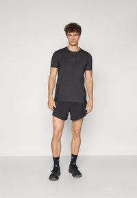 Schwarzes Sport-T-Shirt mit kurzen Ärmeln und Rundhalsausschnitt, mit dezentem Logodetail. Getragen mit schwarzen Shorts und passenden Sportschuhen.