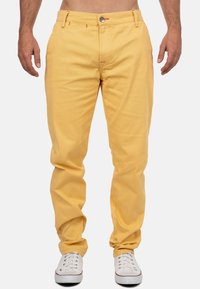 Pantaloni di cotone gialli con vestibilità rilassata, con cuciture arancioni, tasche frontali, chiusura a bottone e indossati con sneakers bianche.