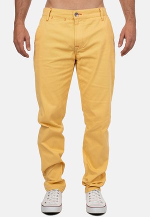 Pantalons en coton jaune avec une coupe décontractée, dotés de coutures orange, de poches avant, d'un bouton de fermeture, et portés avec des baskets blanches.