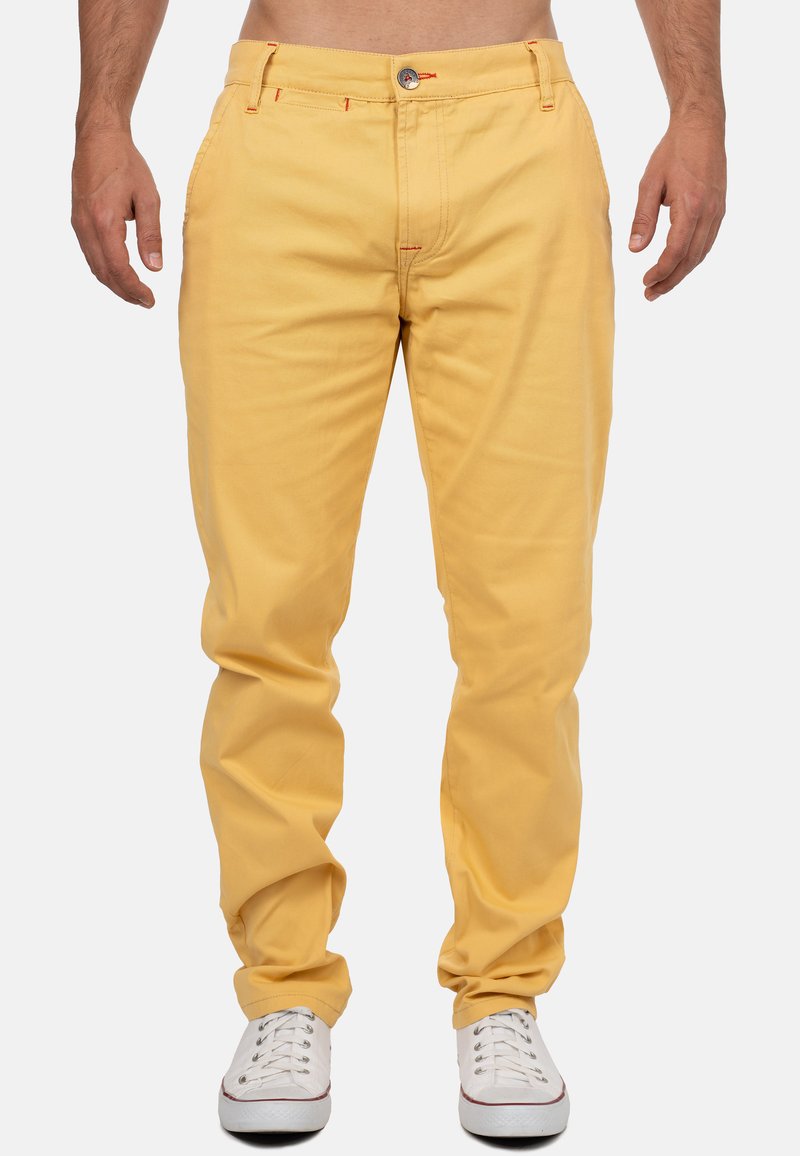 Pantaloni di cotone gialli con vestibilità rilassata, con cuciture arancioni, tasche frontali, chiusura a bottone e indossati con sneakers bianche.