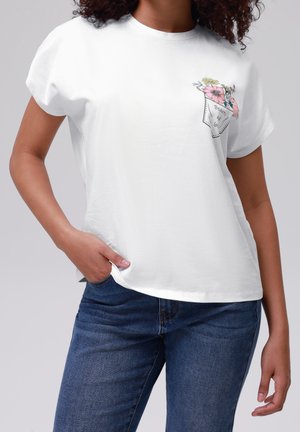 Kobieta w białej koszulce z kwiatowym wzorem na kieszeni i napisem „Piękno miłości”, ubrana w niebieskie dżinsy, z ręką w kieszeni na jednolitym tle.