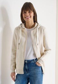 Femme souriante portant un sweat à capuche beige zippé sur un t-shirt blanc et un jean bleu, debout avec une main dans la poche sur un fond clair.