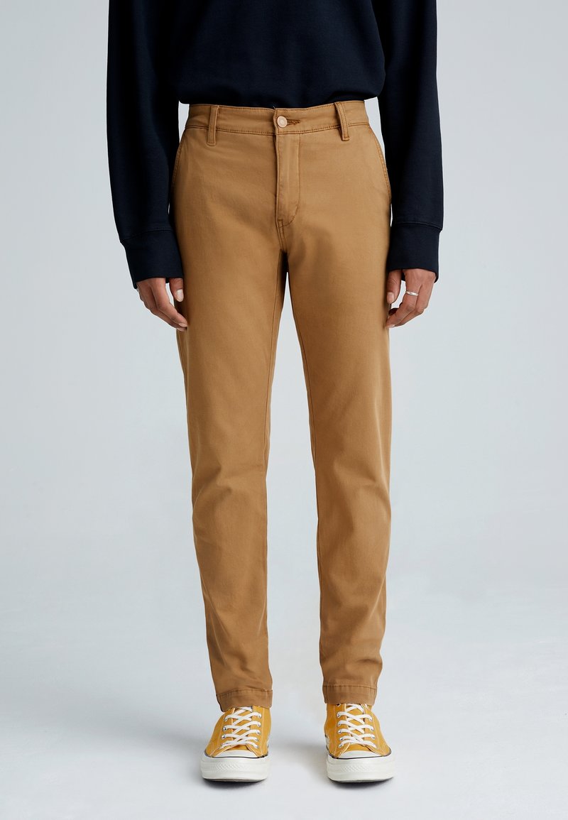Personne portant un pantalon beige coupe droite, un haut noir à manches longues et des baskets jaunes, debout devant un fond gris clair uni.