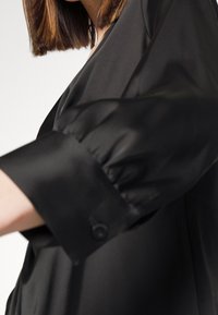 Femme portant un chemisier noir à manches longues bouffantes avec poignets boutonnés, visible de l'épaule à mi-bras sur un fond blanc.