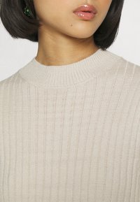 Maglione a costine beige con collo alto e motivo testurizzato. Il tessuto appare morbido con una leggera lucentezza.