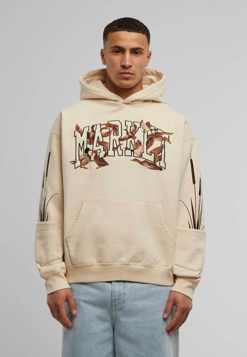 Beige hettegenser med brodert "MARK"-design og fugler. Har kengurulomme og svarte gresslignende detaljer på ermene.