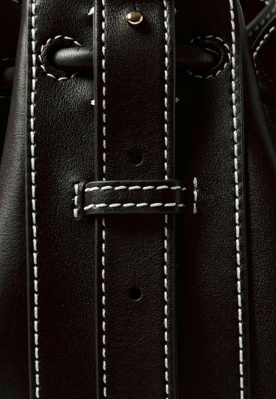 Dettaglio di una borsa in pelle nera con cuciture bianche a contrasto, texture liscia e un accento metallico dorato sulla tracolla.