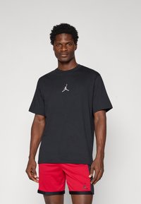Svart bomull t-skjorte med en liten hvit Jumpman-logo på brystet, kombinert med røde shorts med svarte detaljer og logo.
