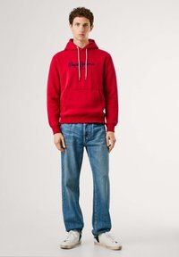 Rode katoenen hoodie met een trekkoordcapuchon, voorzien van een kangoeroe zak en een marineblauw geborduurd logo. Gecombineerd met blauwe denim jeans en sneakers.