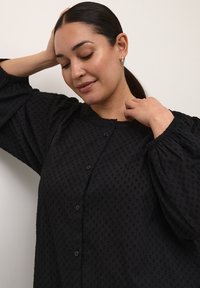 Blusa nera con texture, maniche a sbuffo e scollatura rotonda, caratterizzata da chiusure a bottone e un motivo a pois su tutta la stoffa.