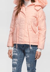 Chaqueta acolchada rosa con capucha, con cierre de cremallera frontal, dos bolsillos laterales y un patrón de acolchado. Combinada con jeans azules desgastados.