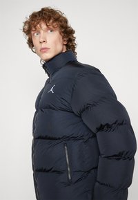 Mornarsko puffer jakna z visokim ovratnikom, horizontalnim prešitim dizajnom in žepi na zadrgo. Ima izrazit logotip na prsih.