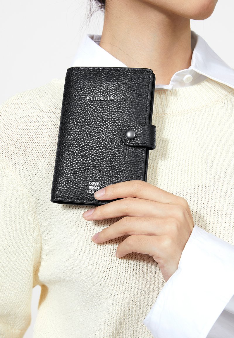 Hånd, der holder en lukket sort læderpung med struktureret overflade, "Victoria Hyde"-logo og trykknap, iført cremefarvet strikket sweater og hvid skjorte.