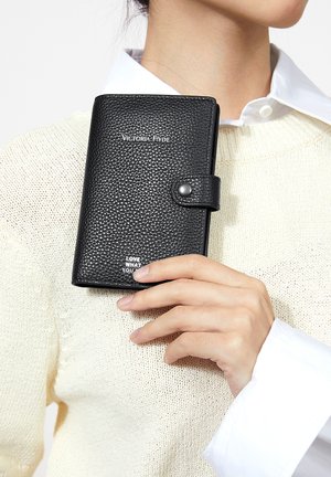 Hand hält geschlossenes, schwarzes, strukturiertes Lederportemonnaie mit "Victoria Hyde"-Logo und Druckknopf, trägt cremefarbenen Strickpullover und weißes Hemd.