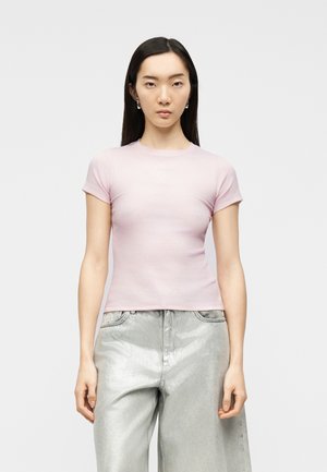 Junge Frau mit langen, dunklen Haaren, die ein hellrosa, figurbetontes Kurzarmshirt und hoch taillierte, silberne, metallisch glänzende Hosen mit weitem Bein trägt.