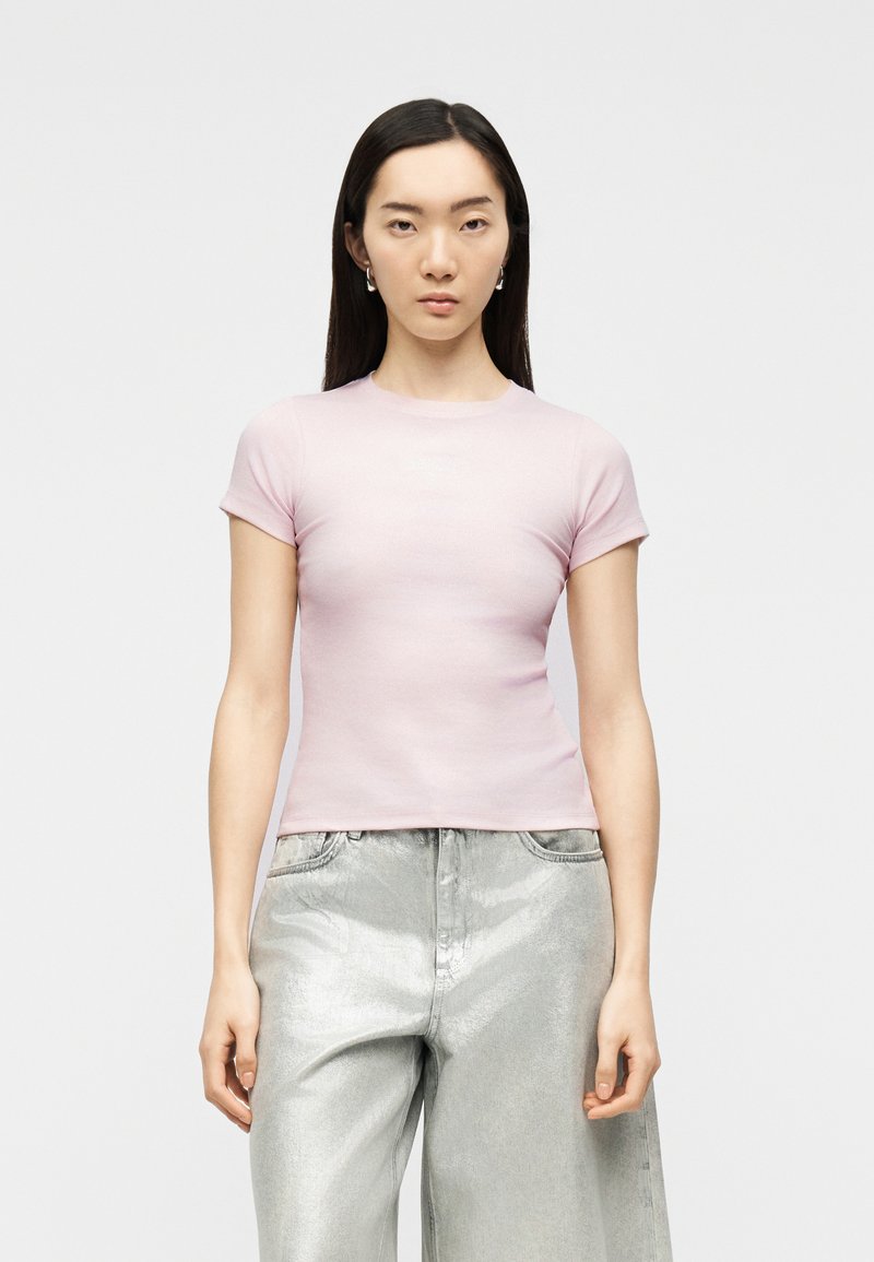 Νεαρή γυναίκα με μακριά σκούρα μαλλιά, φοράει ανοιχτό ροζ εφαρμοστή μπλούζα με κοντά μανίκια και ασημί μεταλλικό παντελόνι με φαρδιά πόδια και ψηλή μέση.