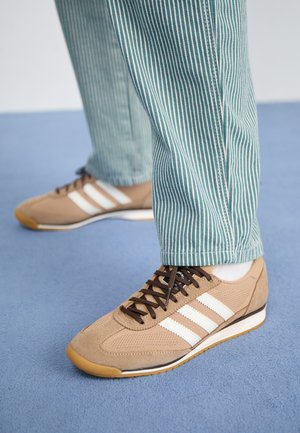 Fødder iført beige sneakers med hvide striber og brune snørebånd, kombineret med grønne og hvide stribede bukser på en blå overflade.