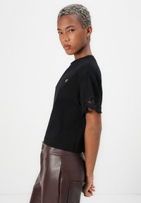 Black cotton t-shirt with a round neckline, short sleeves featuring scalloped lace trim, and a small decorative metallic accent on the chest.