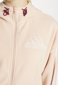 Růžová fleecová mikina na zip s bordó šňůrkami a bílým vyšitým logem Adidas na hrudi. Má vysoký límec.