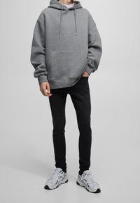 Sudadera gris con un bolsillo frontal, capucha con cordón y puños acanalados, combinada con jeans ajustados negros y zapatillas blancas. Diseño sencillo.