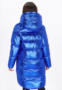 Lang pufferjakke i skinnende blåt materiale, med hætte, quiltet design og elastiske manchetter. Teksturen fremstår blød og reflekterende.